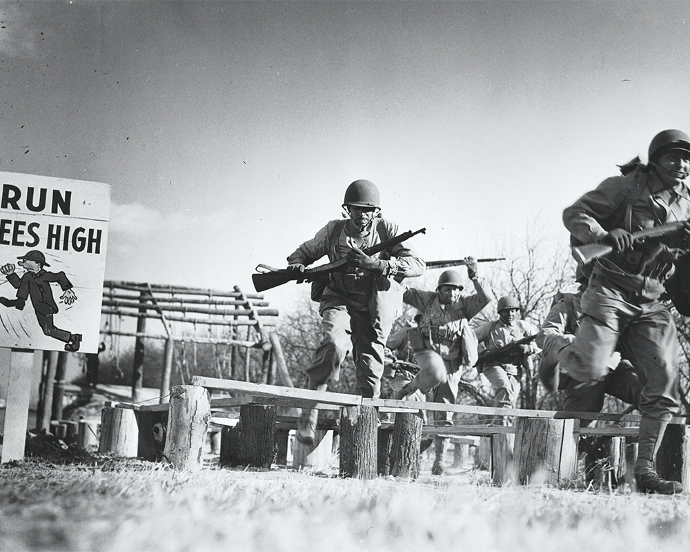 Black & white photo of soldiers running obstacle course with guns