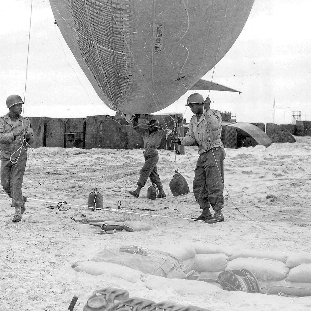 Black & white photo of soldiers setting up huge balloons on the beach