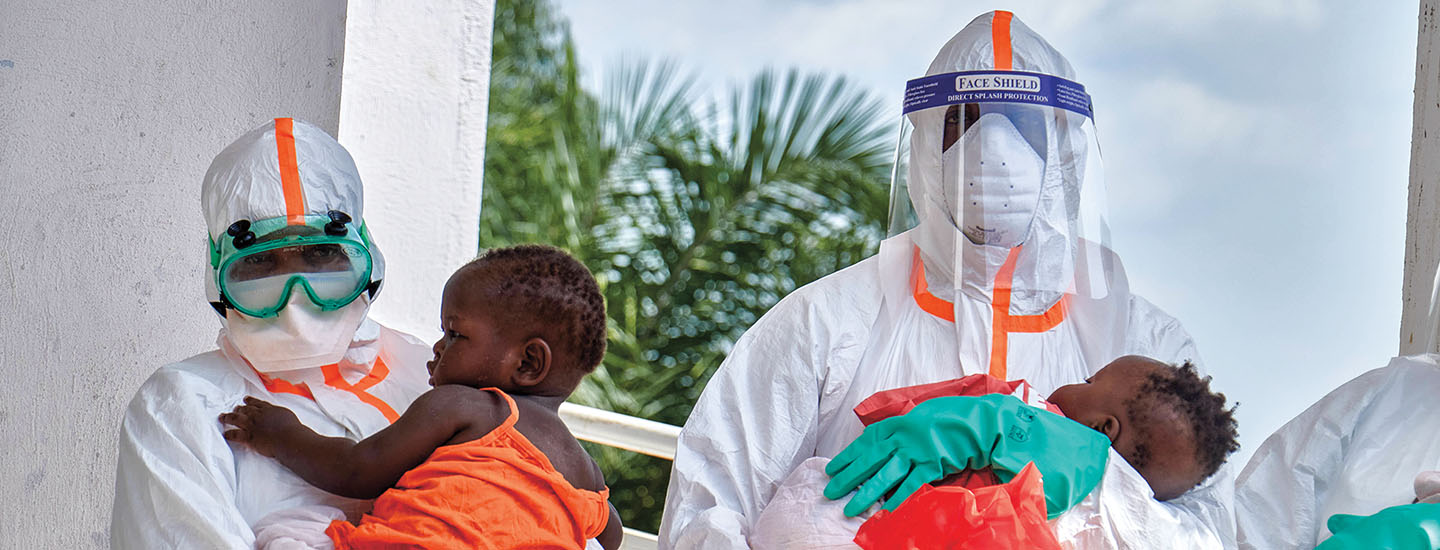 Two healthcare workers in full protective gear each hold a young child outdoors